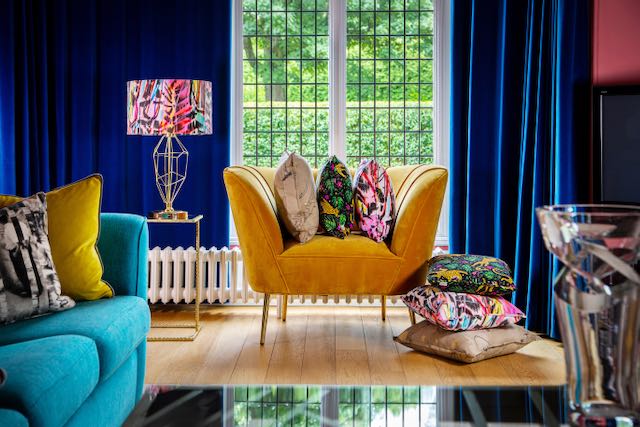 vibrant yellow accent chair piled with patterned cushions in front of a floor to ceiling window. Turquise sofa just seen to the left, more cushions on the floor to the right.