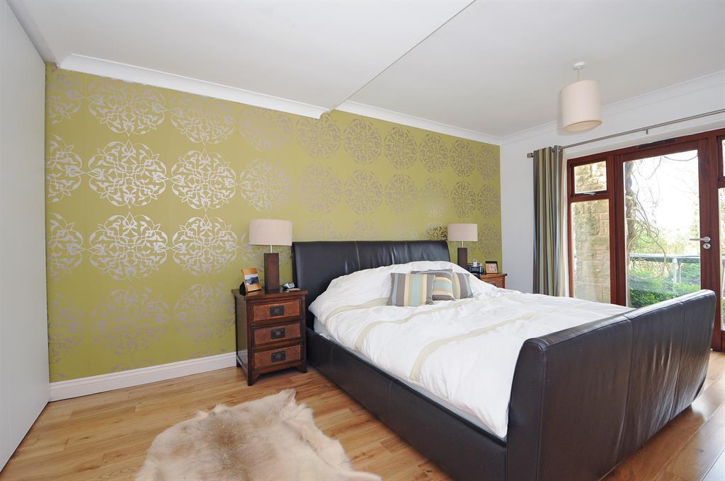 Image of a large modern bedroom. Floor to ceiliing windows on the right, large dark brown sleigh style double bed with white bed linen. Wood floorboards, sheepskin rug and a feature wall behind the bed in a yellow/green colour embossed with large flowers.