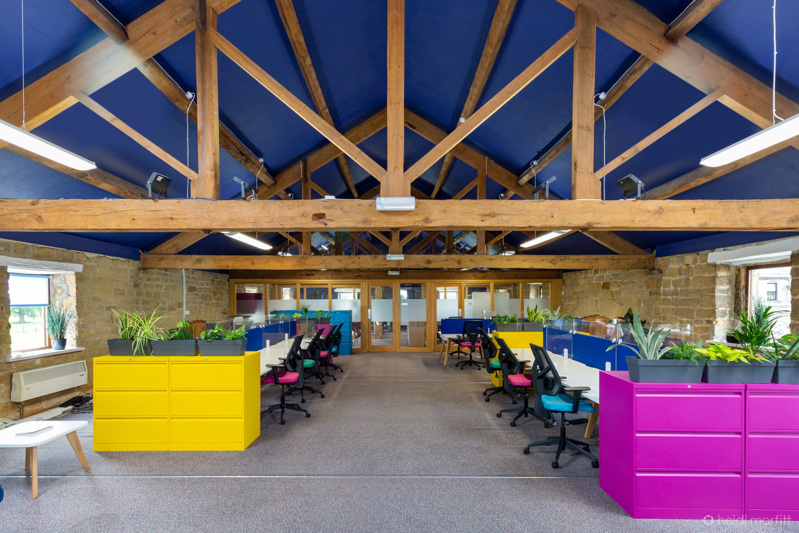 Office in a barn conversion with two banks of desks and office chairs. Lots of colour - a navy ceiling, brightly coloured cabinets and drawers in hot pink and a vibrant yellow