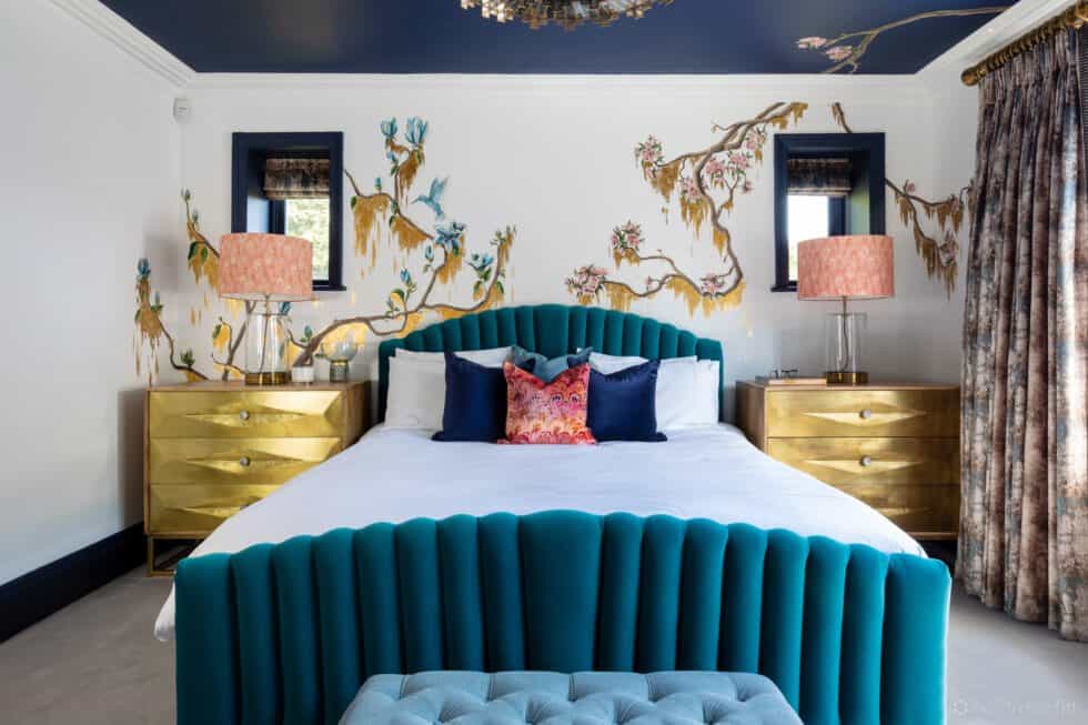 An upholstered teal bed sits against a bespoke mural with gold bedside cabinets and lamps with coral shades