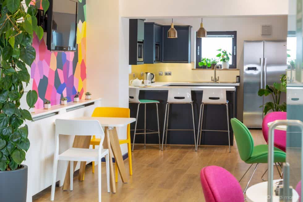 Navy kitchen with gold splash back and white stools at the counter. Vibrant bespoke wallpaper and brightly coloured chairs stand on the oak floor