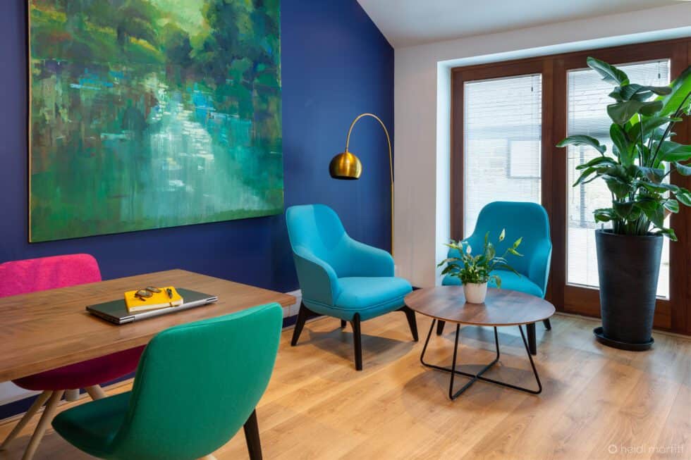Turquoise armchairs provide a break out space in this office with navy walls and bright pink and green chairs at the table