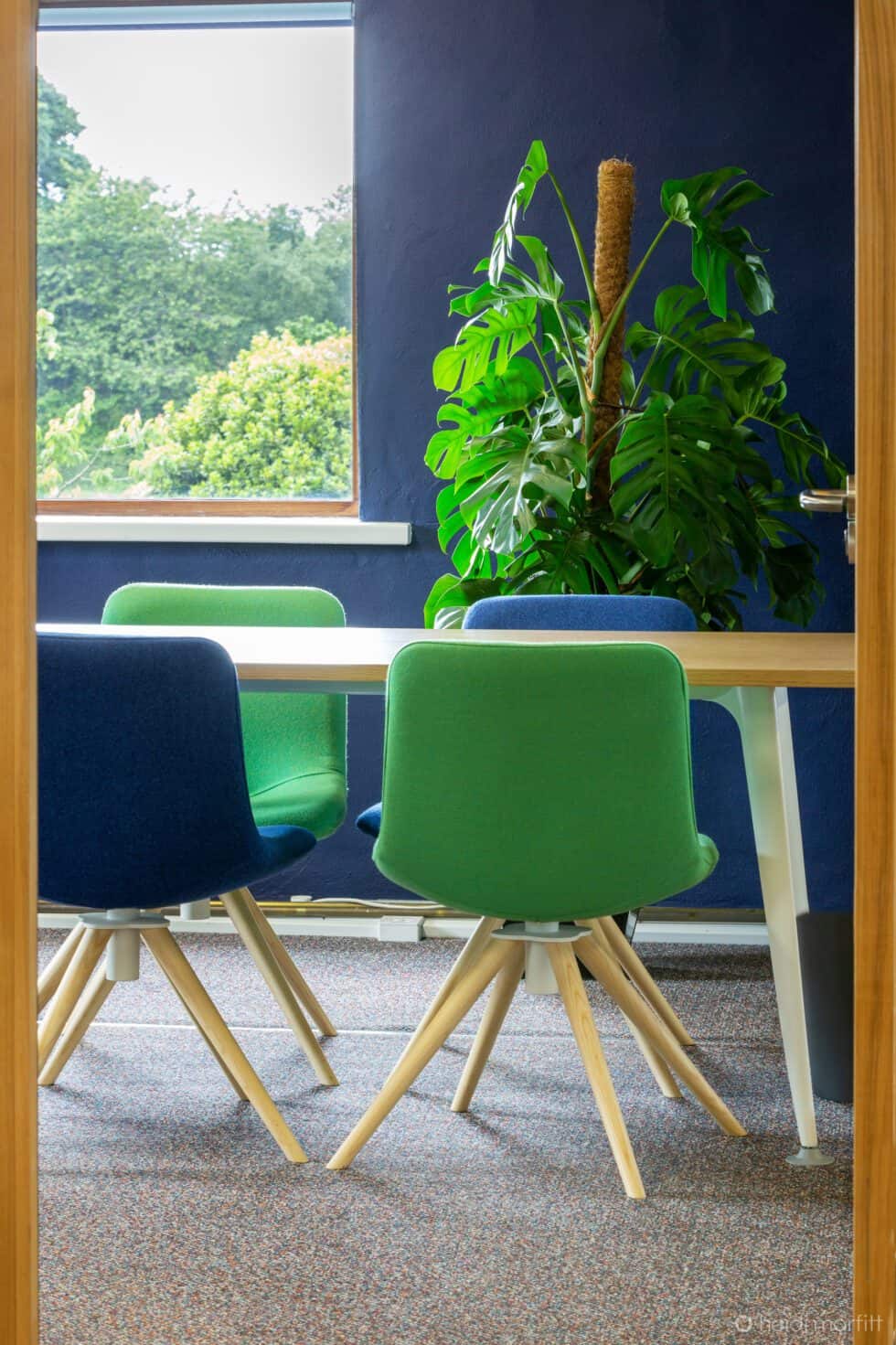 A glimpse through a doorway to an oak and white desk with green and blue chairs