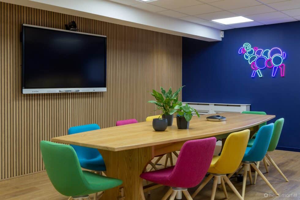 Meeting room with wooden panelling board table and vibrantly coloured chairs. Neon light logo sits on the navy walls.