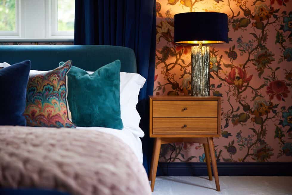 Pink patterned wallpaper provides pattern to this master bedroom where a chestnut bedside table sits next to a teal bed with blue and patterned cushions