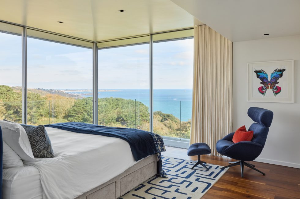 Blue and white bedroom with bespoke Jennifer Manners rug and navy armchair with large picture windows overlooking the sea